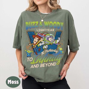 May include: Moss green t-shirt featuring a graphic of Buzz Lightyear and Woody from Toy Story. The design includes the text "Buzz & Woody Lightyear" and "To Infinity and Beyond". The shirt is a relaxed fit.