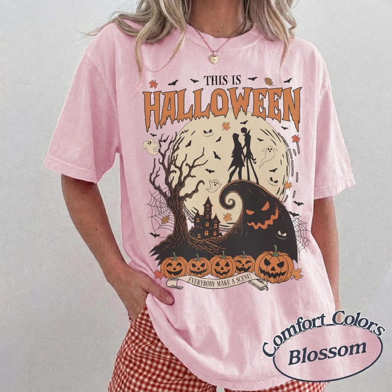 May include: Pink t-shirt with a Halloween-themed graphic. The design features a full moon, silhouettes of characters, a haunted house, pumpkins, and the text "THIS IS HALLOWEEN" in orange. The shirt also has the text "EVERYBODY MAKE A SCENE".
