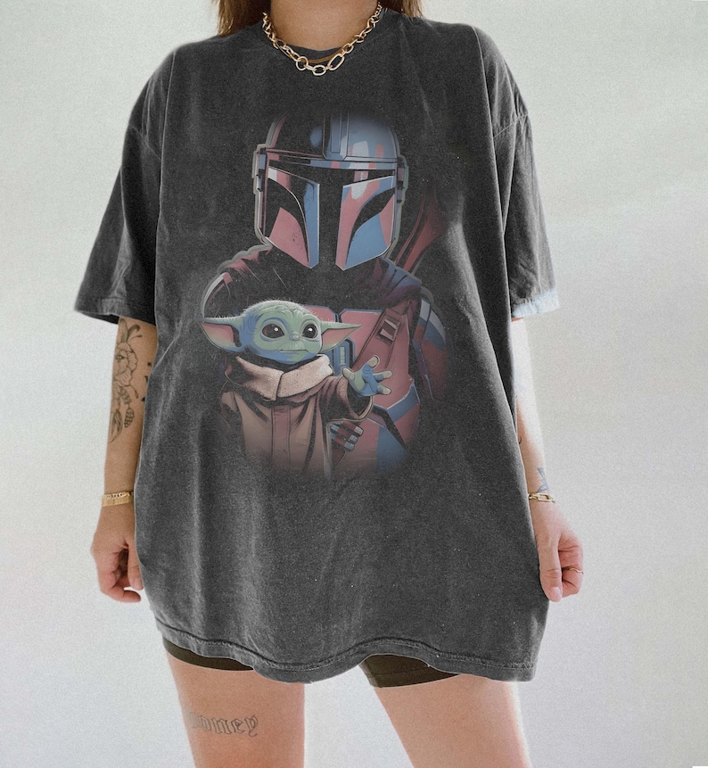 May include: A charcoal gray short-sleeve t-shirt featuring a graphic print of a helmeted warrior in silver and blue armor standing behind a small green creature with large ears wearing a tan robe, with the creature reaching out one hand.