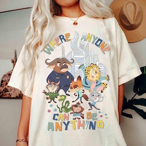 Puede incluir: Camiseta de color crema con un gr&aacute;fico colorido de varios animales de dibujos animados y el texto "WHERE ANYONE CAN BE ANYTHING". El dise&ntilde;o incluye un tel&oacute;n de fondo de paisaje urbano y acentos florales, creando una est&eacute;tica l&uacute;dica y vibrante.