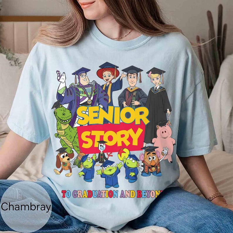 K&ouml;nnte beinhalten: Hellblaues T-Shirt mit einem farbenfrohen Aufdruck von Toy Story-Charakteren in Abschlusskleidung. Der Text "SENIOR STORY" ist in Gelb und Rot dargestellt, darunter "TO GRADUATION AND BEYOND". Das Shirt besteht aus einem weichen Material.