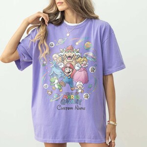 Puede incluir: Una camiseta de color lila con un gr&aacute;fico colorido de personajes de Super Mario Galaxy. El dise&ntilde;o incluye a Mario, la Princesa Peach, Bowser y otros personajes, con el texto "The Super Mario Galaxy" y "Custom Name".