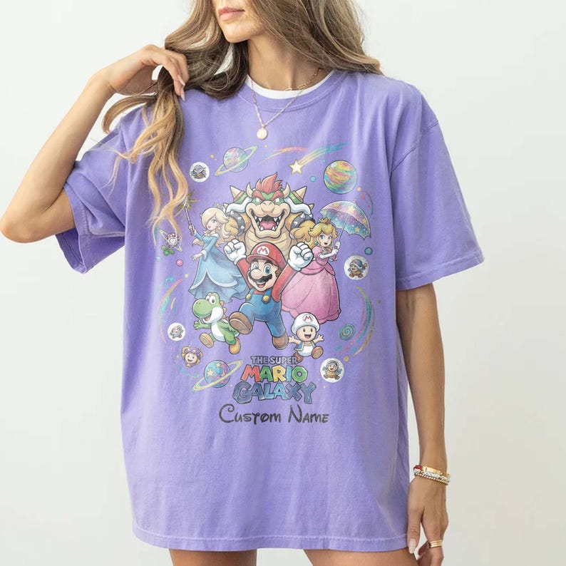 Puede incluir: Una camiseta de color lila con un gr&aacute;fico colorido de personajes de Super Mario Galaxy. El dise&ntilde;o incluye a Mario, la Princesa Peach, Bowser y otros personajes, con el texto "The Super Mario Galaxy" y "Custom Name".