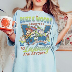 May include: Light blue t-shirt featuring Buzz Lightyear and Woody from Toy Story. The graphic includes the text "Buzz & Woody Lightyear" and "To Infinity and Beyond". The shirt has a relaxed fit and short sleeves.