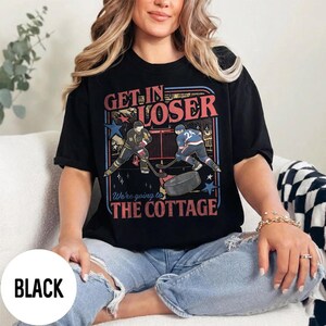 May include: Black t-shirt with a hockey-themed graphic. The design features two hockey players, a puck, and the text "Get in Loser" and "We're going to the Cottage" in red and blue.