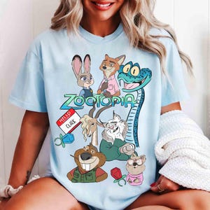 May include: Light blue t-shirt featuring characters from the animated film "Zootopia." The design includes Judy Hopps, Nick Wilde, and other characters, with the film's title in green and blue. A "Hello, Clark" name tag is also visible.