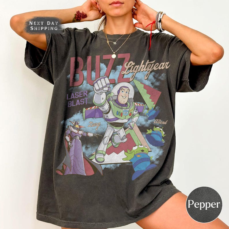 May include: A charcoal gray oversized t-shirt featuring a graphic print of Buzz Lightyear, Zurg, and green aliens. The text reads BUZZ Lightyear, LASER BLAST, Zurg, and Aliens. The shirt has short sleeves and a crew neckline with a vintage-style wash.