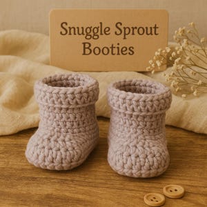 May include: A pair of light purple crochet baby booties with rolled cuffs. The text "Snuggle Sprout Booties" is on a brown card behind the booties. Two small wooden buttons are at the bottom.