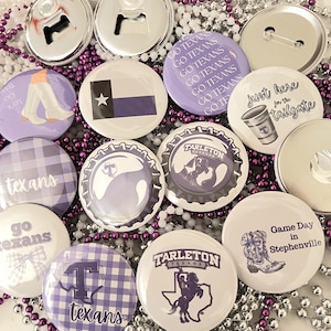 May include: A collection of round buttons and bottle openers in shades of purple and silver. The buttons feature various designs, including the Texas flag, cowboy boots, and the words "Go Texans" and "Game Day in Stephenville."