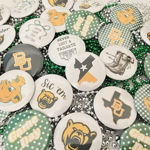 May include: A collection of round buttons with various designs, including the Baylor University logo, bear illustrations, and phrases like "Sic 'em" and "Game Day." The buttons are white, green, gold, and gray, with a green beaded necklace.