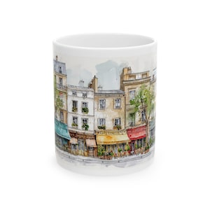 May include: White ceramic mug with a watercolour illustration of a European street scene. The artwork shows buildings with colourful awnings, shops, and trees. The mug has a standard handle and a glossy finish.
