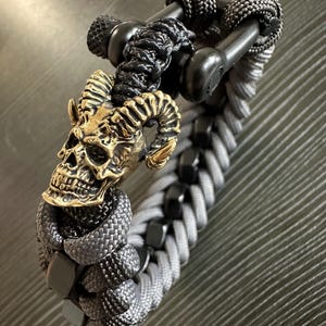May include: A gray and black paracord bracelet featuring a brass skull with ram horns. The bracelet has a braided design with black metal accents and a black clasp. The skull is detailed with intricate carvings.