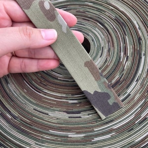 May include: A close-up of a roll of camouflage fabric with a strip of the same fabric held in a hand. The fabric features a pattern of green, brown, and white, suitable for outdoor gear and apparel.