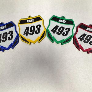 May include: Four colorful racing number plate ornaments. Each plate is a different color: blue, yellow, green, and red. Each plate has the number "493" in large black font and the word "NAME" above the number.