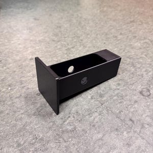 May include: A black metal bracket with a rectangular body and a vertical plate. The bracket has two circular holes and a small, recessed area. The surface has a textured finish.