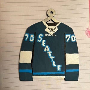 Puede incluir: Adorno de jersey de hockey de madera pintado a mano en tonos azul, blanco y negro. El jersey presenta la palabra "SEATTLE" en blanco, el número "70" y la marca "BAUER". El adorno cuelga de una percha plateada.