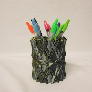May include: A 3D-printed pen holder with a geometric design in shades of black and green. The holder is filled with various colored pens, including orange, blue, pink, green, and purple. The pen holder is a desk organizer.