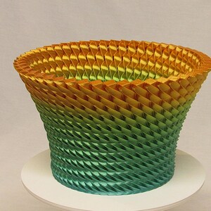 May include: A decorative, gradient-colored basket. The basket transitions from a golden-yellow hue at the top to a vibrant green at the base. The design features a repeating pattern of angled, layered elements, creating a textured surface.