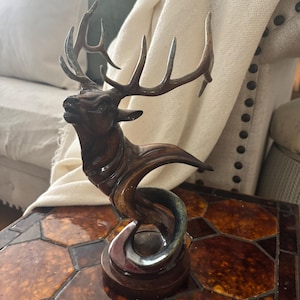 May include: A bronze-colored elk sculpture with large antlers. The elk is posed in a dynamic, upward-flowing design, with a swirling base. The sculpture is placed on a table with a brown and orange tiled surface.