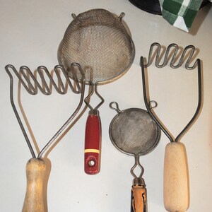 May include: Four vintage kitchen tools: two potato mashers with wooden handles, one with a red handle, and a small strainer with a black handle. The strainer is round and has a mesh design. The potato mashers have a wire design.