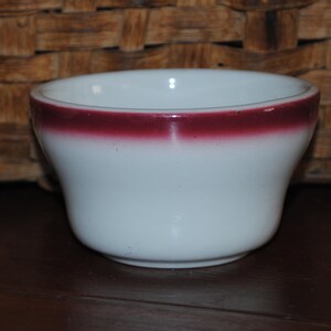 May include: A white ceramic bowl with a single red band around the rim. The bowl is slightly flared at the top and has a rounded bottom.