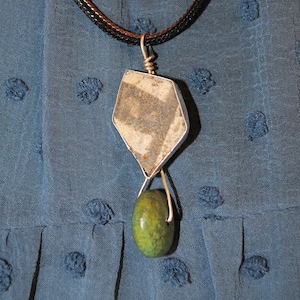 May include: A silver pendant necklace with a gray and white stone and a green stone hanging from it. The necklace is on a blue fabric background.