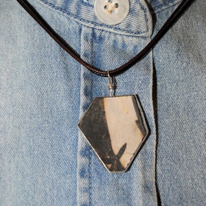 May include: A silver pendant necklace with a hexagonal shaped stone pendant. The stone is a mix of black and white with a rough texture. The necklace is on a denim shirt.