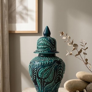 May include: A teal ceramic jar with a matching lid, featuring a raised relief pattern of swirling teardrop shapes and small dots. The vessel has a bulbous body, a tapered neck, and a pointed finial on the lid, finished in a glossy glaze.