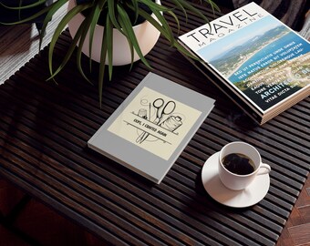 Op de afbeelding: Een grijze notitieboek met de tekst "OOPS, I CRAFTED AGAIN" en een illustratie met naaithema. Een kopje koffie, een tijdschrift en een potplant staan ook op de donkere houten tafel.