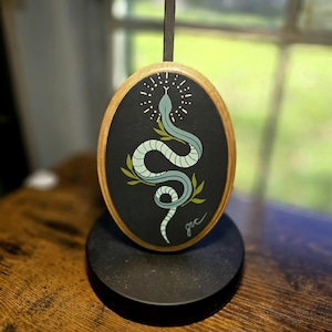 May include: An oval wooden plaque with a natural wood edge features a black painted center. A light blue snake is coiled around green leaves, topped with a white sunburst design. The initials gvc are painted in white at the bottom right.
