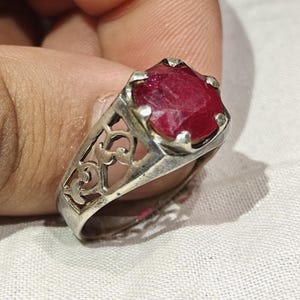 May include: A silver ring with an ornate filigree design and a large, faceted, deep red gemstone. The ring is held up against a white background, showcasing the intricate metalwork and the vibrant color of the stone.