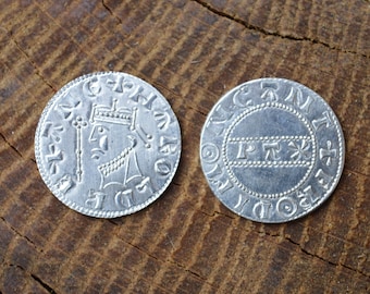 May include: Two silver coins with detailed engravings. One coin features a profile of a crowned figure holding a scepter. The other coin has a central design with text. Both coins are set against a textured, brown wooden surface.