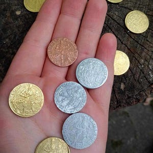 Puede incluir: Colección de monedas antiguas en varios acabados metálicos, incluyendo oro, plata y cobre. Cada moneda presenta diseños en relieve detallados de figuras e inscripciones. Las monedas se exhiben sobre una superficie de madera, mostrando su estética histórica.