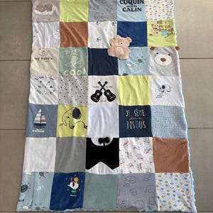 Peut inclure: Une couverture patchwork faite à partir de vêtements pour bébés, avec une variété de couleurs et de motifs. La couverture comprend des carrés avec du texte, des graphiques d'animaux et un petit ours en peluche. La couverture est posée sur un sol carrelé.