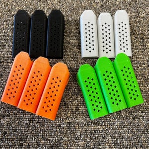 May include: A selection of rectangular objects in black, white, orange, and green, each with a pattern of small square holes. The items are displayed on a textured, brown surface.