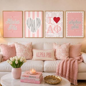 Op de afbeelding: Vier ingelijste wandkunstprints met Alpha Phi-branding in tinten roze, wit en grijs. De ontwerpen bevatten streeppatronen, een hartvorm, een rood hartpictogram en geschulpte randen, weergegeven boven een witte bank met roze kussens.