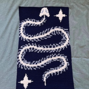 Könnte beinhalten: Ein marineblau gehäkeltes Wandbehang mit einem weißen und hellblauen Schlangenmuster und zwei weißen Sternformen. Das handgefertigte Textilkunstwerk ist rechteckig und hat ein strukturiertes Aussehen.