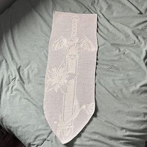 Pode incluir: Uma tapeçaria de parede de crochê creme com um design detalhado de uma espada com asas e detalhes florais. A peça feita à mão é exibida em uma superfície verde clara, mostrando o intrincado padrão vazado.
