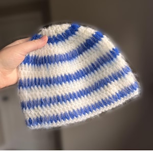 Könnte beinhalten: Eine gestrickte Mütze mit horizontalen Streifen in Weiß und Blau. Die Mütze ist aus weichem, strukturiertem Garn gefertigt. Die Streifen sind gleichmäßig angeordnet. Ein klassischer Stil, ideal für kaltes Wetter, dieses Accessoire ist gemütlich.
