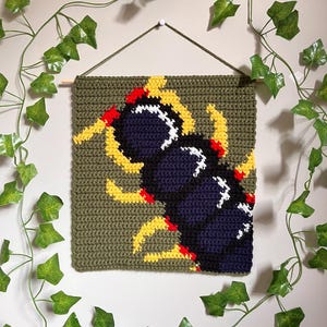Peut inclure: Suspension murale carrée en crochet vert olive représentant un mille-pattes stylisé. Le mille-pattes est principalement bleu marine avec des accents blancs, jaunes et rouges. Accroché à une baguette en bois et une ficelle.