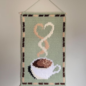 May include: A crocheted wall hanging depicting a coffee cup with steam forming a heart shape. The design uses white, brown, and beige on a green backdrop, framed with a brown and beige border. It's hung from a wooden dowel, creating a cosy feel.