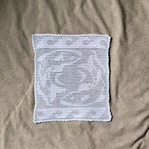 May include: A square, light blue crocheted doily with a detailed openwork design. The edges are finished with a scalloped border. The doily is placed on a neutral-toned, textured fabric.