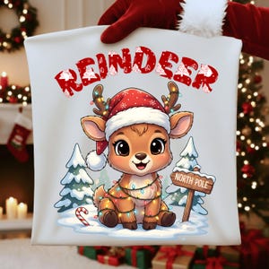 Op de afbeelding: Een wit kussen met een kerstontwerp. Het toont een cartoon rendier met een kerstmuts en lichtsnoer, met het woord "REINDEER" erboven. Een bordje leest "NORTH POLE".