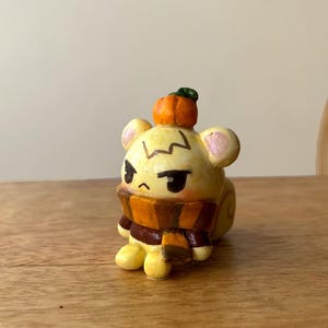 Puede incluir: Figura amarilla pintada a mano de un personaje de dibujos animados con una calabaza en la cabeza y una bufanda. El personaje tiene una expresión de mal humor y está sentado sobre una superficie de madera. La bufanda es marrón y naranja.