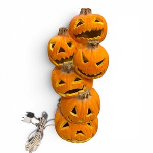 May include: A stack of six orange, carved pumpkin decorations with various facial expressions. The pumpkins are lit from within and have a power cord with a plug attached. The pumpkins are stacked vertically, creating a Halloween-themed decoration.