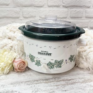 May include: White Rival Crock-Pot slow cooker with a clear lid and dark green trim. The slow cooker has a floral design with the words "RIVAL CROCK-POT" printed on the front. It has a white control knob with settings for Off, Low, and High.