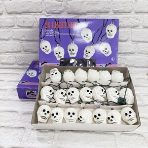 May include: A string of 20 white skull-shaped lights, displayed in an open box with a purple box in the background. The purple box reads "20 LIGHT SET". The lights are connected by black wires, perfect for Halloween decorations.