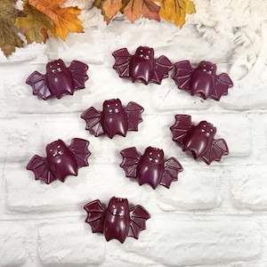 May include: Seven dark purple bat-shaped beads arranged in a scattered pattern. Each bead features a smooth, rounded body, outstretched wings with defined segments, and two small white fangs visible on the face of each individual bat figure.