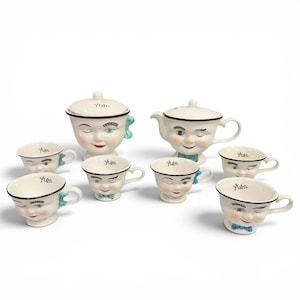 May include: A ceramic tea set featuring a teapot, a lidded sugar bowl, and six teacups, all designed with cream-colored faces, winking eyes, red lips, and the word "Yum." printed on the rims. Each piece includes a small handle and colorful accents.