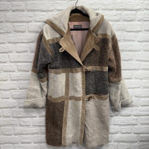 May include: A patchwork coat in shades of brown, gray, and beige, with a hood and toggle closures. The coat features suede accents and a textured fabric, hanging on a black hanger against a white brick wall. The label inside the coat reads "Jinx".